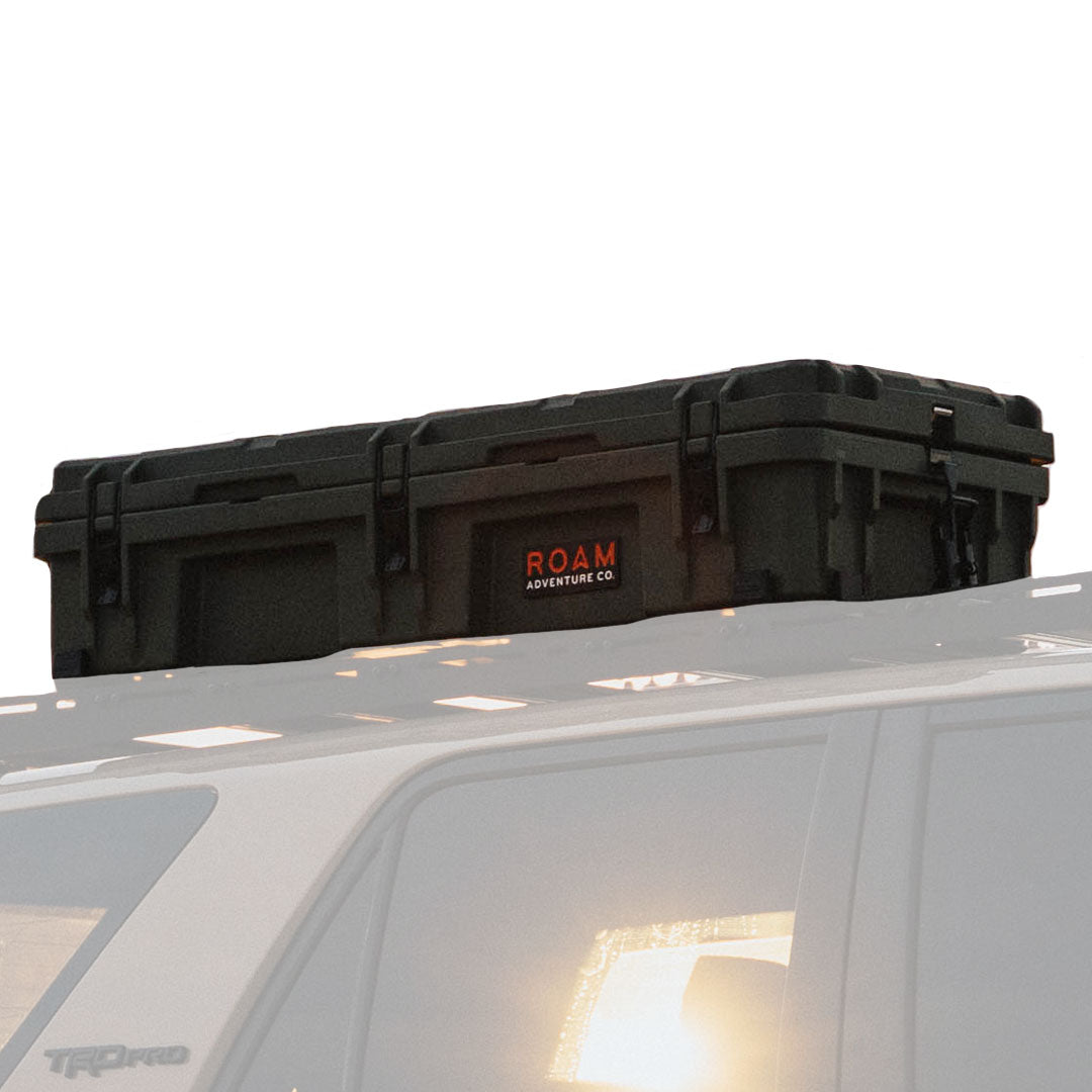 A black ROAM Adventure Co. hard-sided storage case mounted on the roof rack of a white vehicle, shown against a bright white sky.