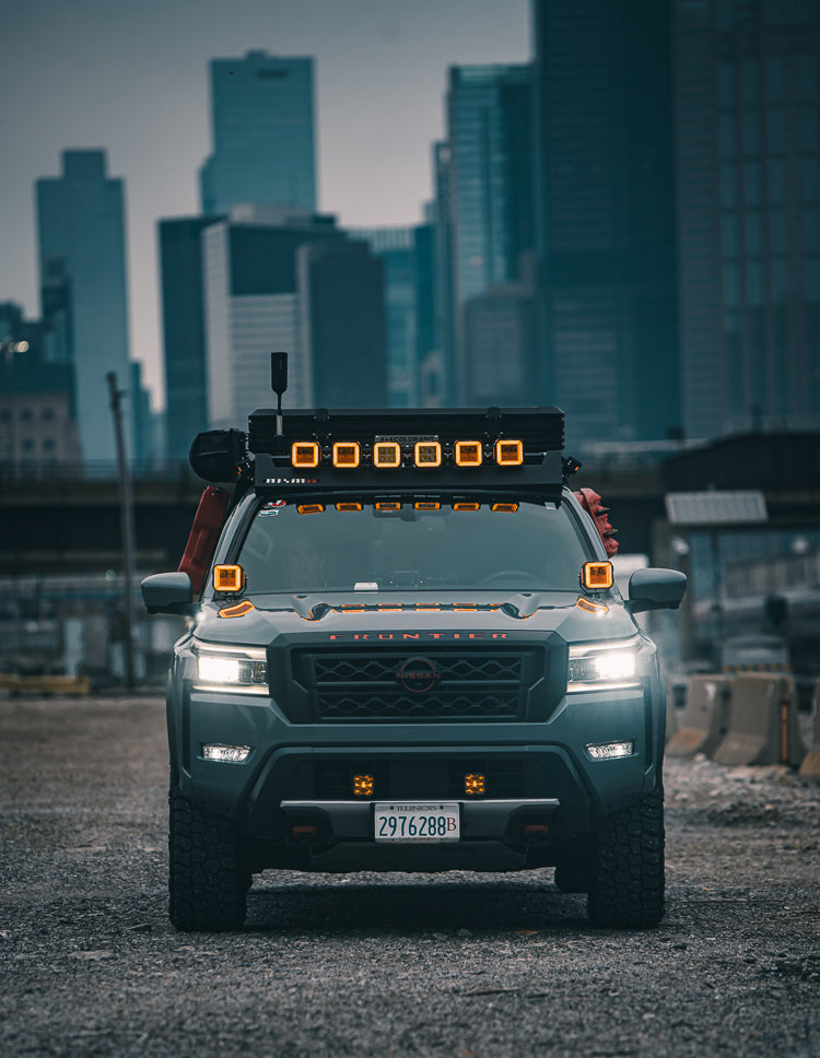 A dark gray Nissan pickup truck with an off-road roof light bar and auxiliary lights, parked on a gravel surface with a blurred city skyline in the background.