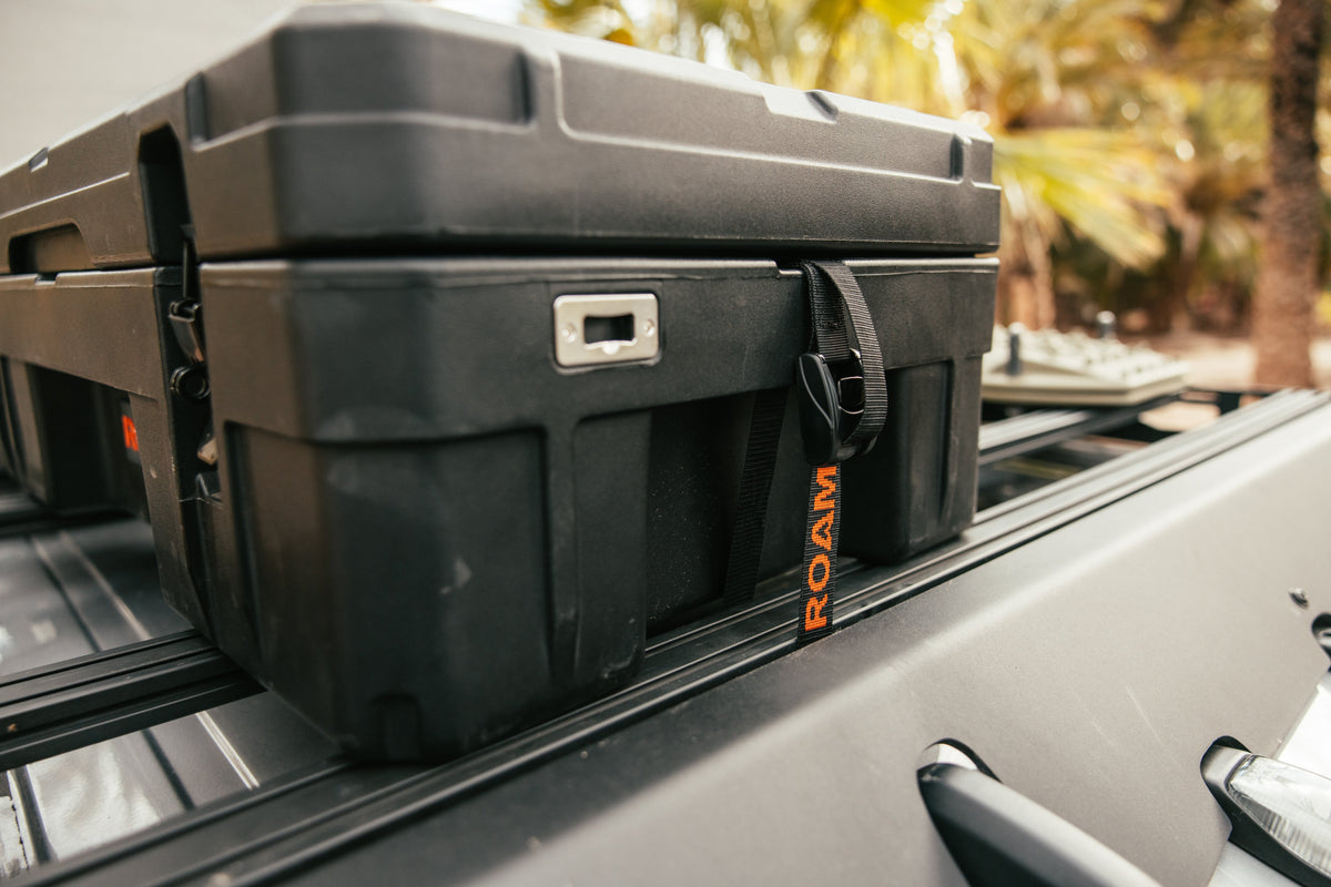 Black ROAM hard-shell storage case on a car roof rack with blurred natural background