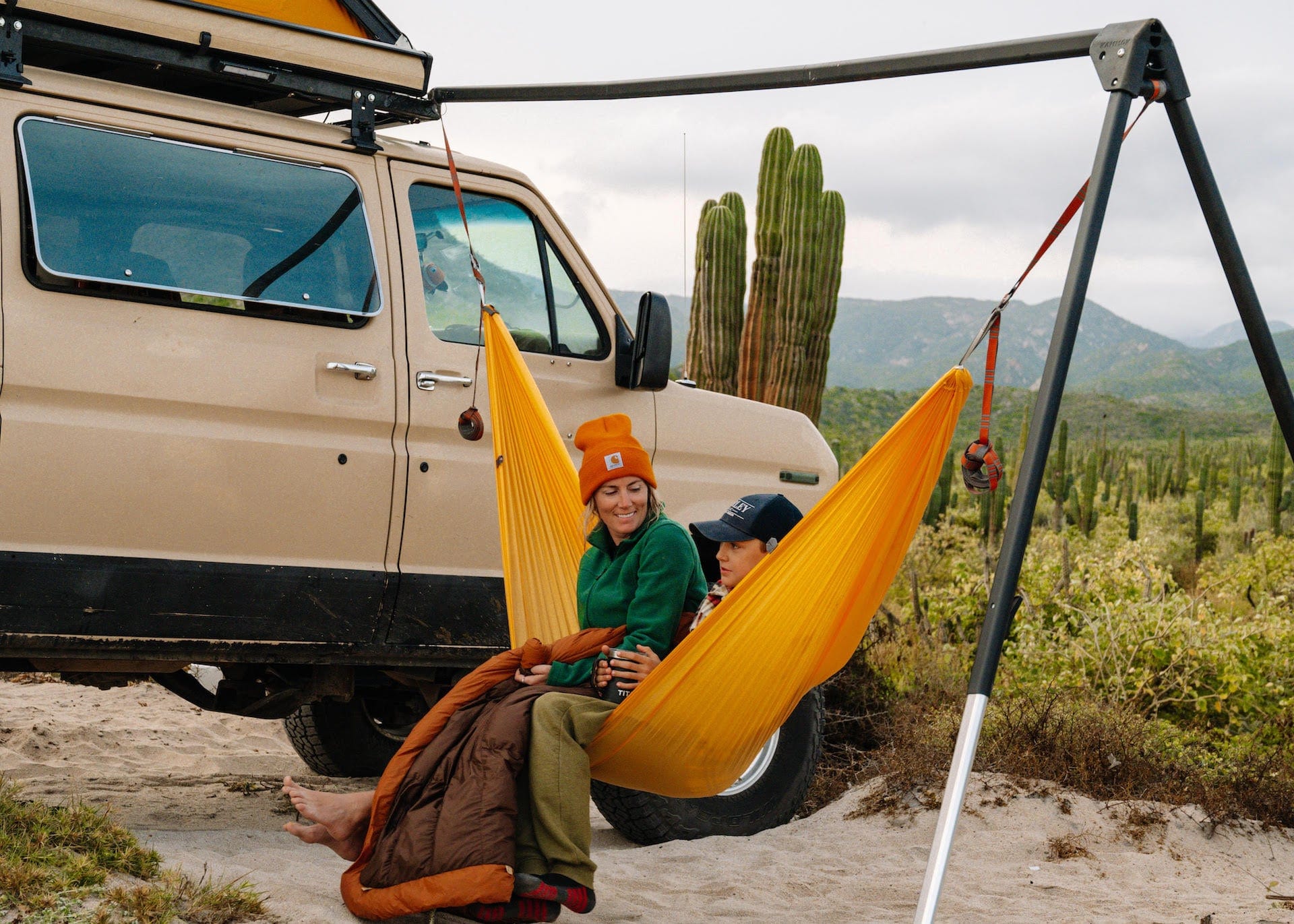 Two people in a yellow hammock in front of a van with cacti and mountains in the background.