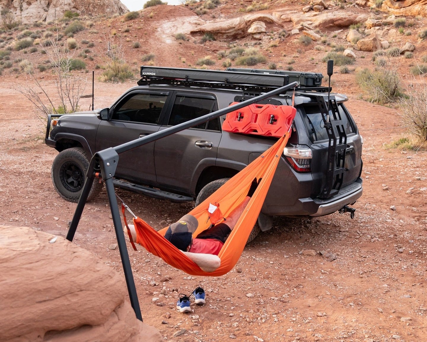 Person in an orange hammock attached to a roof rack on a desert landscape with a vehicle.