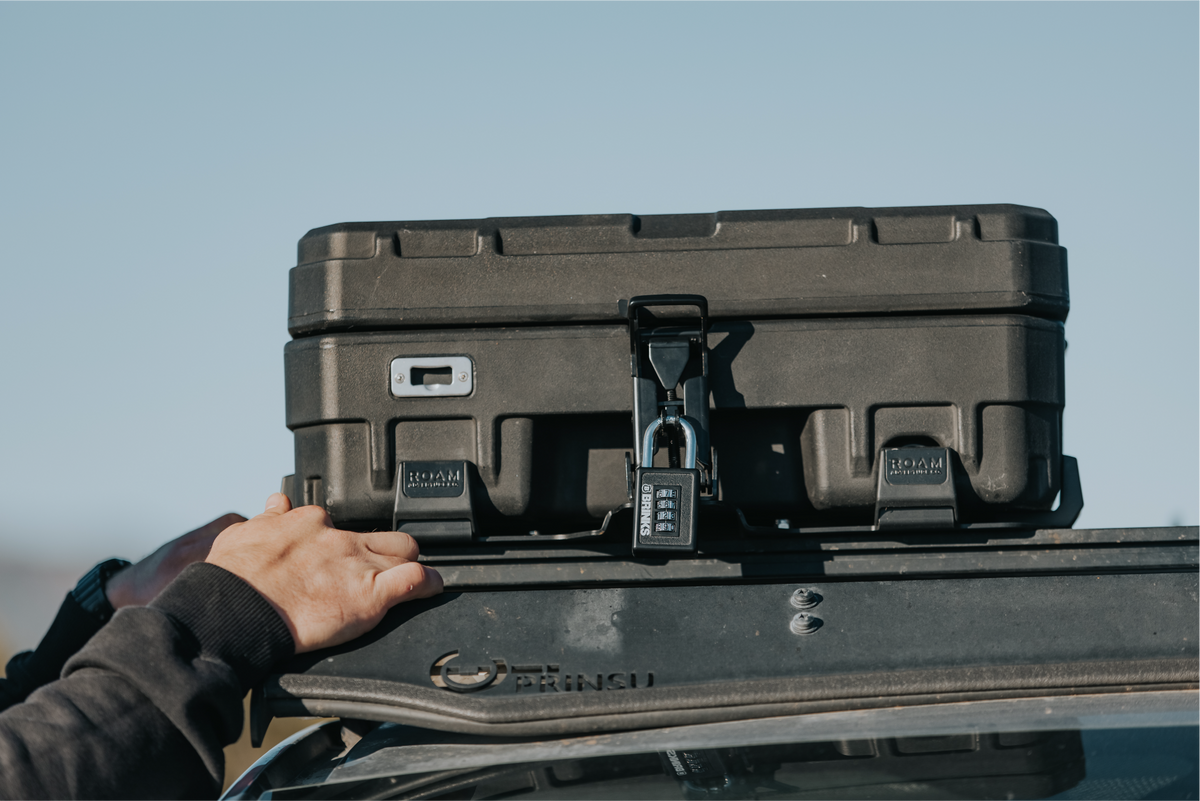 Person securing a black rooftop storage box on a vehicle with a clear blue sky background