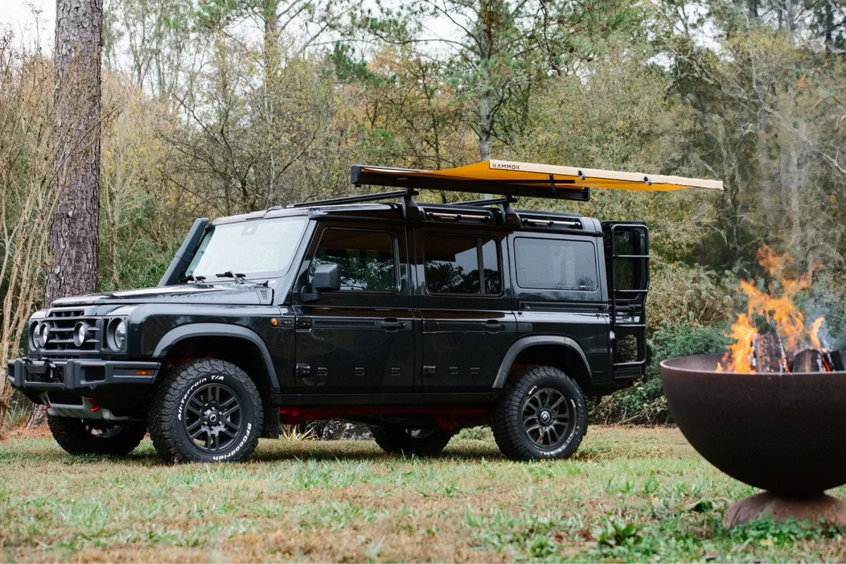 Black SUV with a roof rack in a forest setting near a fire pit.