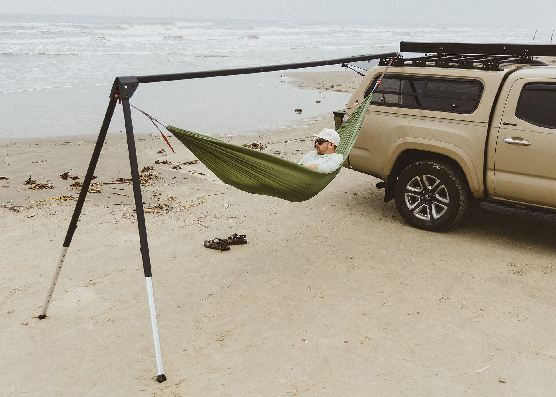 Person in a green hammock attached to a car on a sandy beach.
