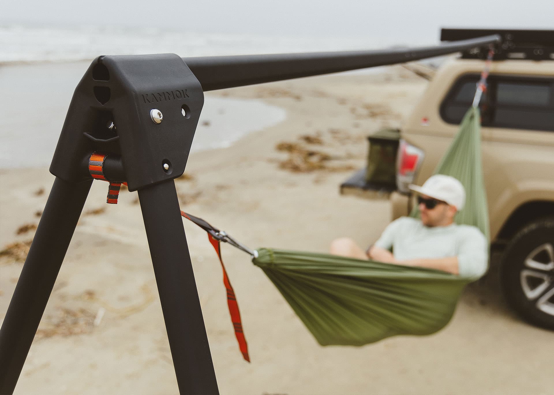 Hammock attached to a roof rack with a person relaxing in it, parked vehicle in the background.