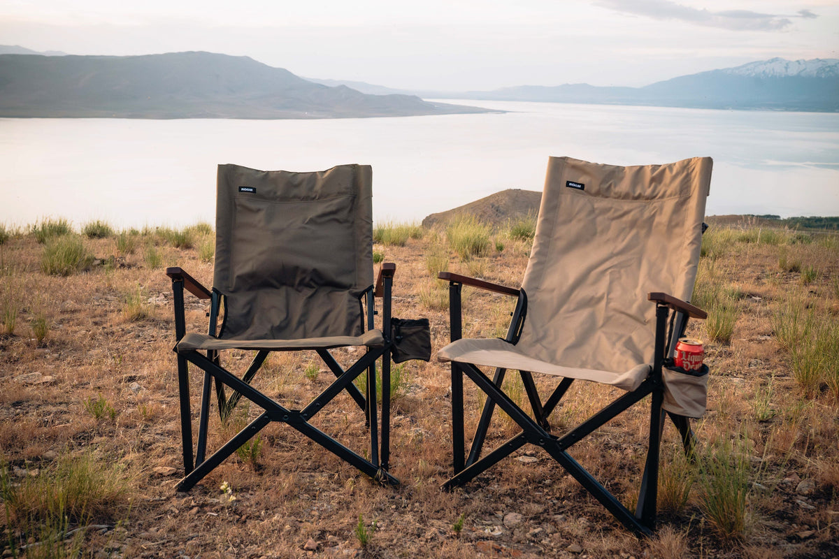 Two camping chairs on a grassy field with a scenic background of mountains and water.