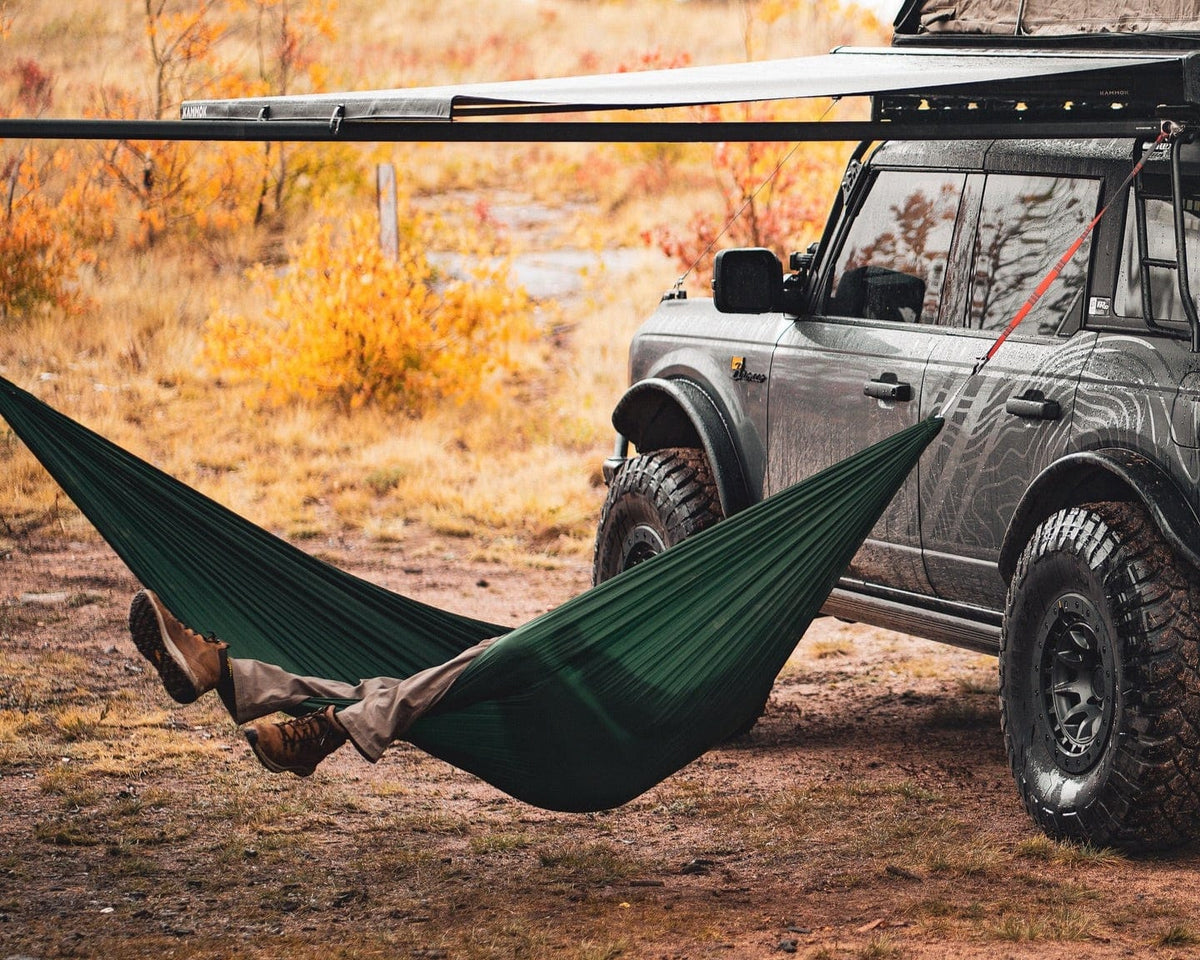 Person relaxing in a green hammock attached to a vehicle in a natural setting